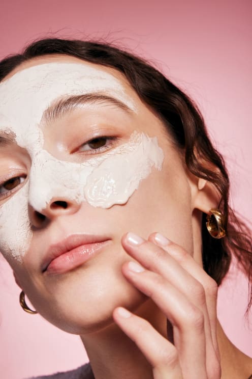 Woman With White Clay On Her Face Gently Touches Her Chin Against A Pink Background