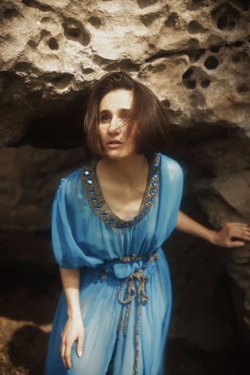 A Woman In A Flowing Blue Dress Crouches Near A Rocky Cave Entrance, Looking Ahead