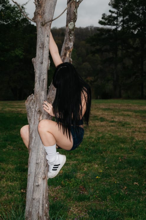 A Girl With Long Black Hair Climbs A Tree In A Grassy Park