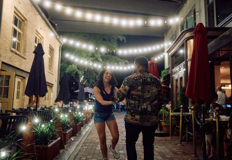 Couple Smiles And Dances Under Hanging Lights On Narrow Street Between Outdoor Restaurants At Night