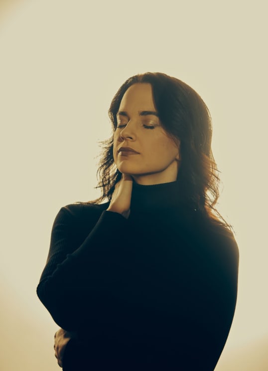 A Darkhaired Woman Dressed In Black With Her Hand Against Her Neck