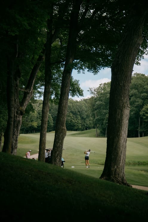 A Female Golfer Swings A Club On A Treelined Golf Course While Other Golfers Look On