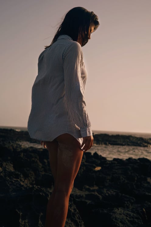 Woman In Brown Hair Bikini And Striped Shirt Standing On Rocky Shore Near Sea At Sunset