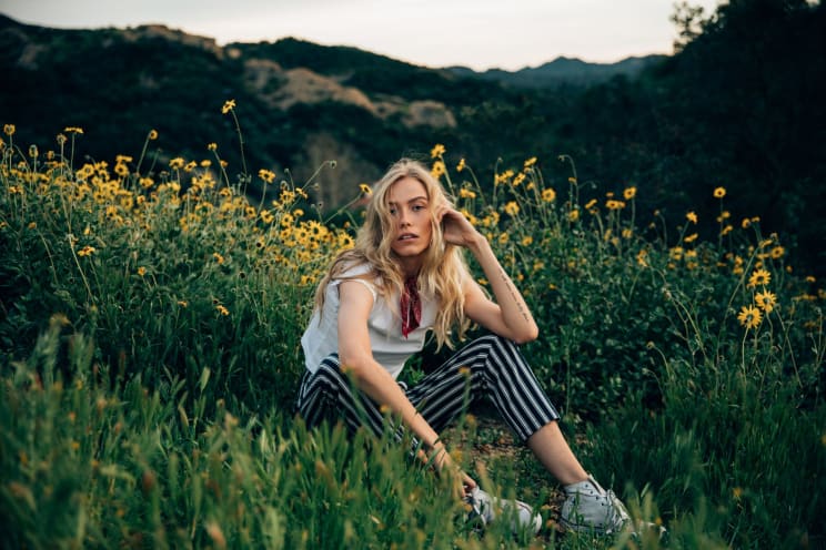 A Blonde Teenage Girl Sitting Among Vegetation With A Neutral Expression
