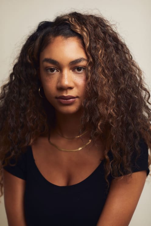 A Young Woman Wearing A Double Necklace And Blue Shirt Poses For A Photograph