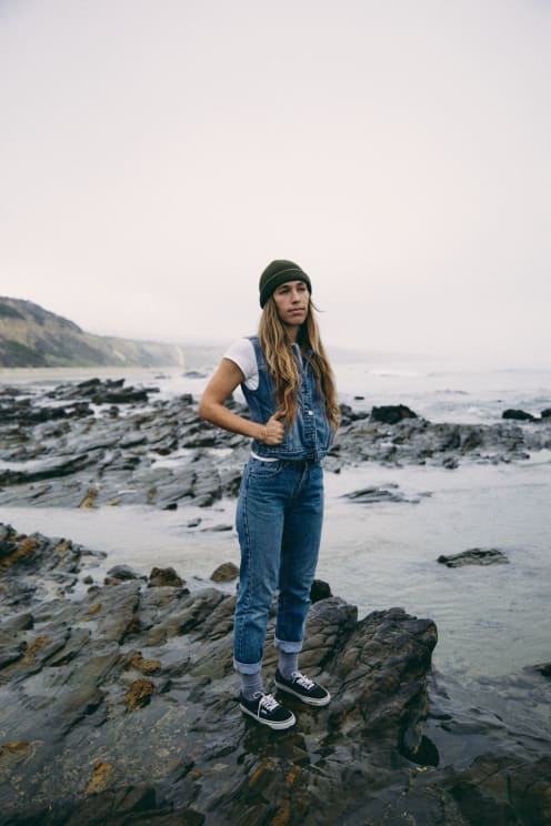 Woman With Hat Denim Vest And Jeans Stands On Rock On The Coast