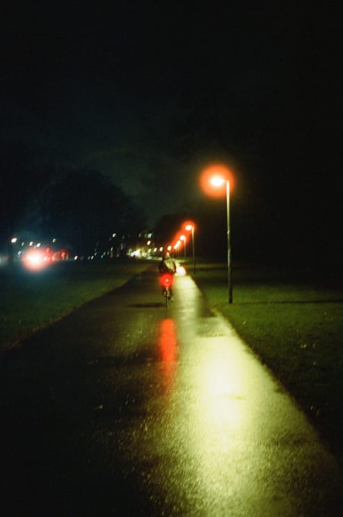 Cyclist Rides Slick Bike Path At Night Light Reflects Off Wet Pavement Car Parked To Left