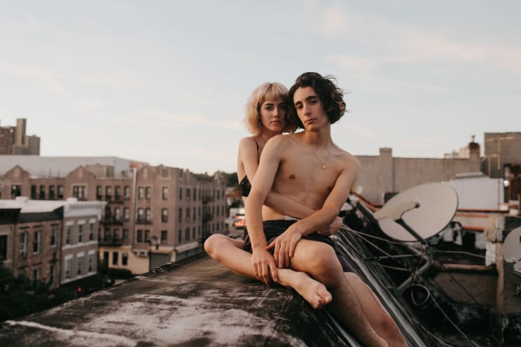 Young Shirtless Couple Sitting On The Roof Of A Building In The City At Evening