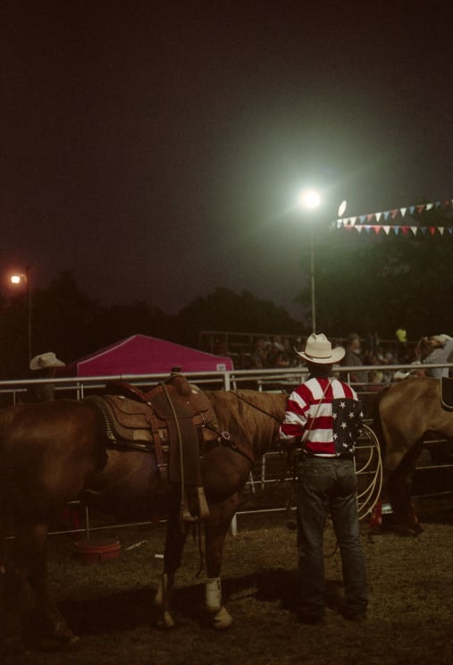 A Cowboy In An American Flag Shirt And White Hat Waits With His Horse At A Nighttime Rodeo Show