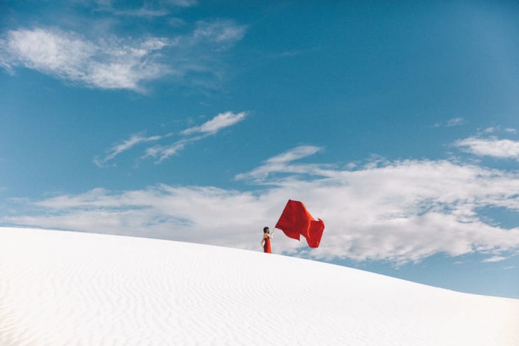 A Woman In A Red Dress Stands On A White Sand Hill Holding A Red Flag Under A Bright Blue Sky With White Clouds