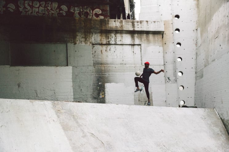 A Man Outside A Worn City Building Juggles A Soccer Ball With His Knee