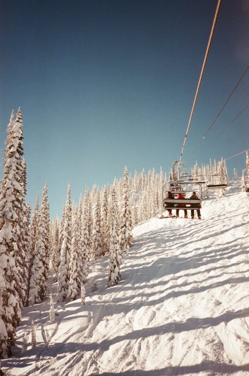 Three People Ride A Ski Lift Up The Side Of A Mountain In British Columbia During Winter
