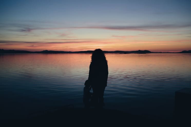A Woman With Curly Hair And A Toddler Girl Standing In Water Are Silhouetted Against The Sky At Sunset