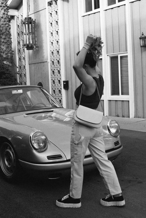 A Woman Walks Towards The Passenger Side Of An Old Convertible As She Puts Up Her Hair