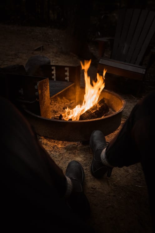 A Man's Feet In Front Of An Outdoor Fire In A Metal Ring In A Campsite At Night