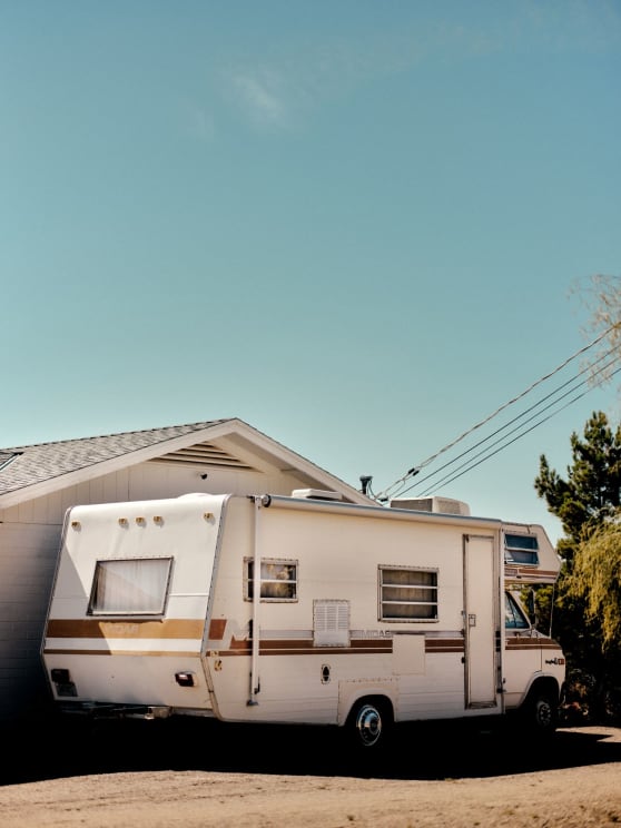 A Vintage 1970s White Camper, Parked By A House On A Dirt Road On A Sunny Day