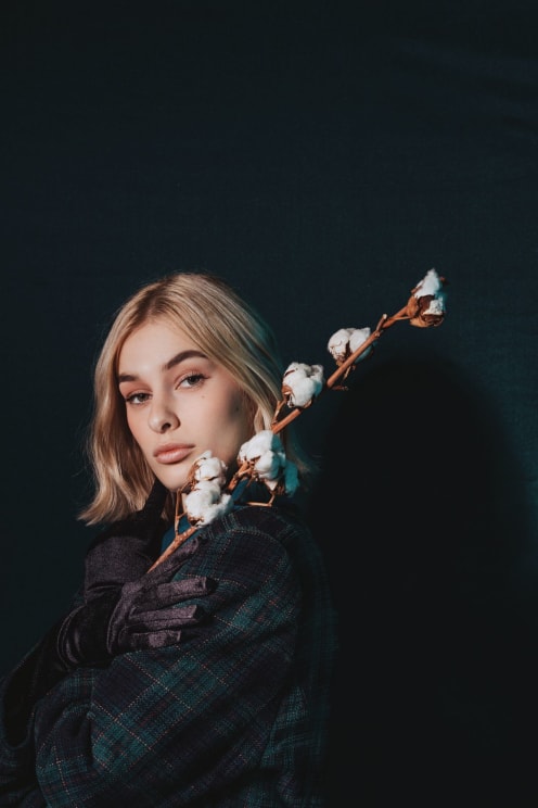 A Young, Blonde Woman Wearing A Plaid Jacket Poses While Holding A Stick Of Cotton Over Her Shoulder