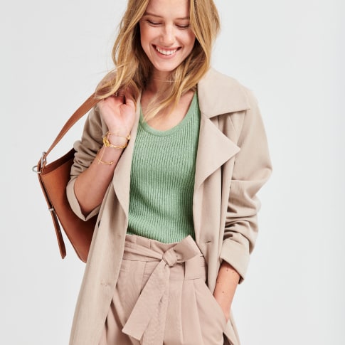 Stitch Fix model wearing a petite camel trench coat, petite camel tapered trousers, a green top and gold bracelets and a brown leather bag.