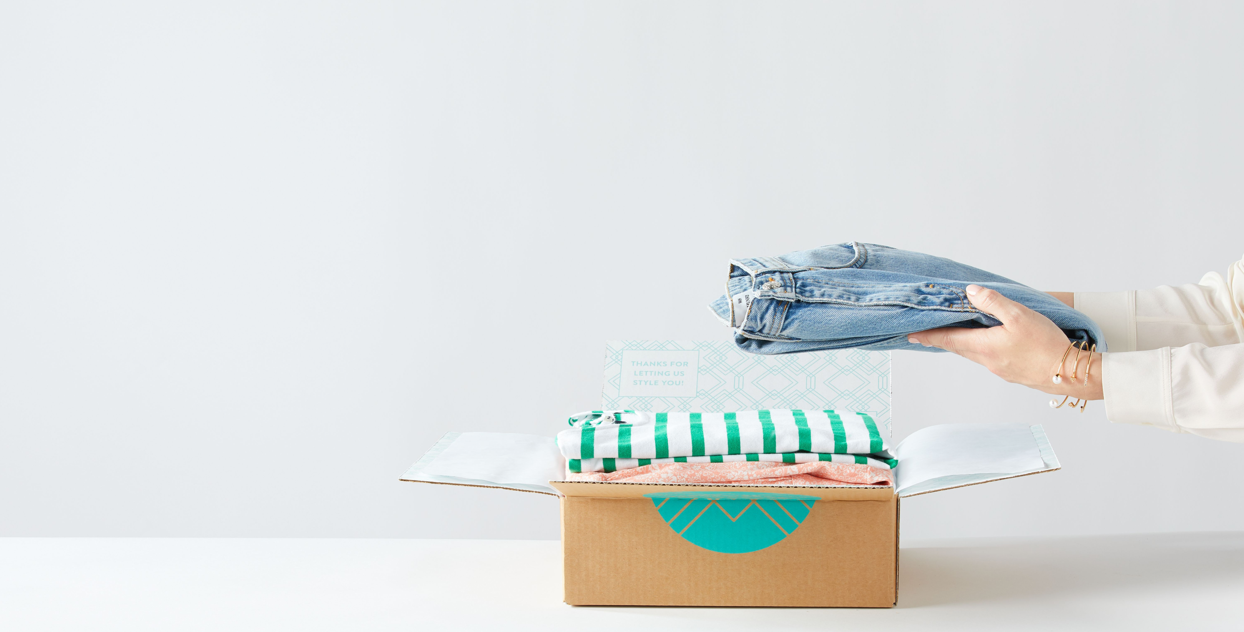 Stitch Fix stylist packing a box. Female hands with the visible sleeves of a white shirt and gold bangle on the left wrist, lifting a pair of Stitch Fix womens blue jeans out of a Stitch Fix box containing other female clothing, including a green breton stripe top and pale pink floral dress.