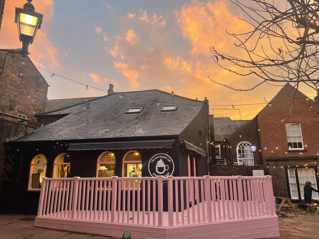 The Buttered Cat's black-fronted cafe and pink terrace in Kinmont Arcade under an orange sunset sky, strung with festoon lights.
