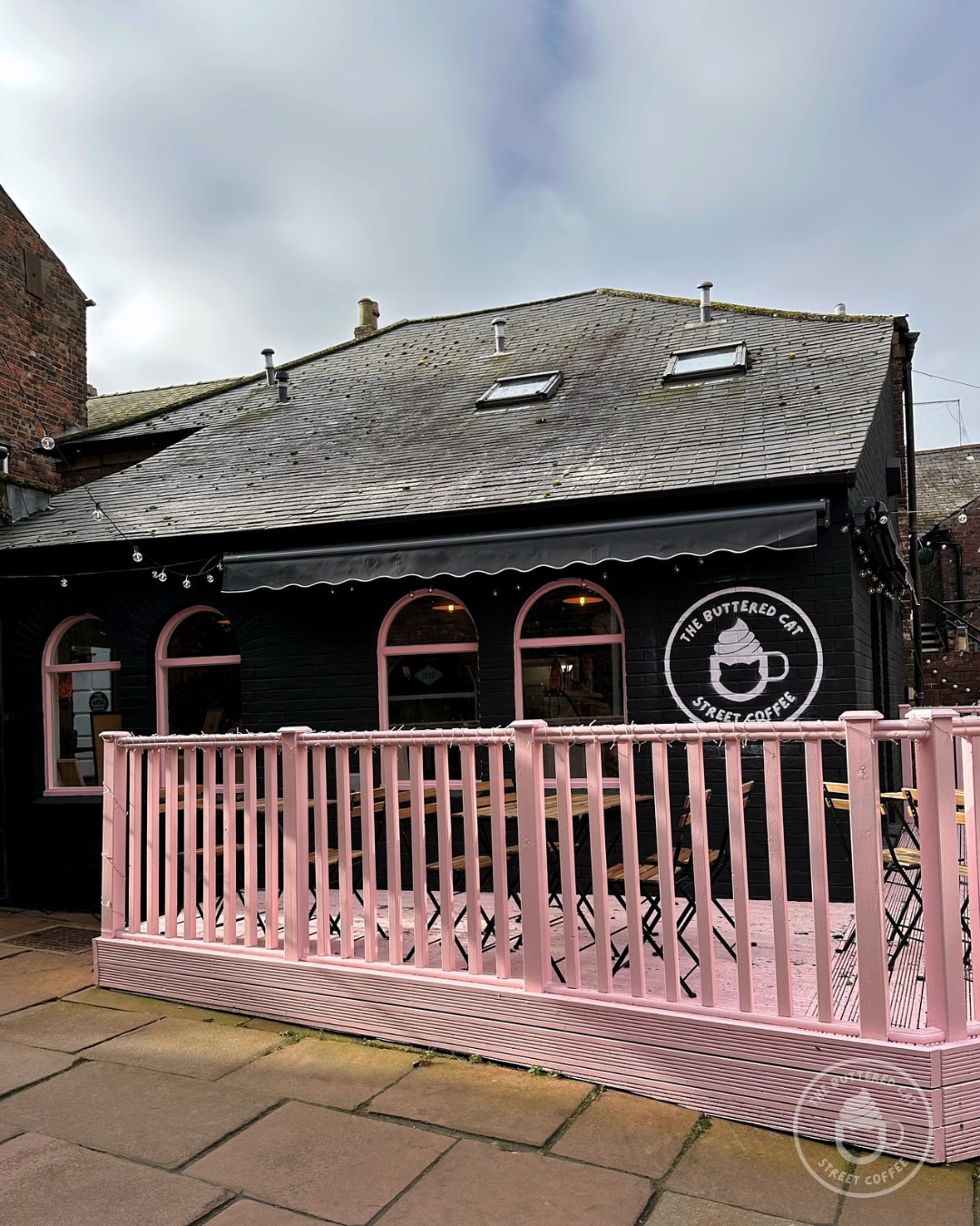 The cafe's three pink arched windows and awning seen from the arcade courtyard, with the painted logo on the black brick wall.