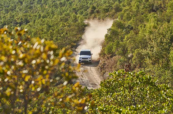 Un Discovery blanc roule sur une route de campagne poussiéreuse dans les bois.