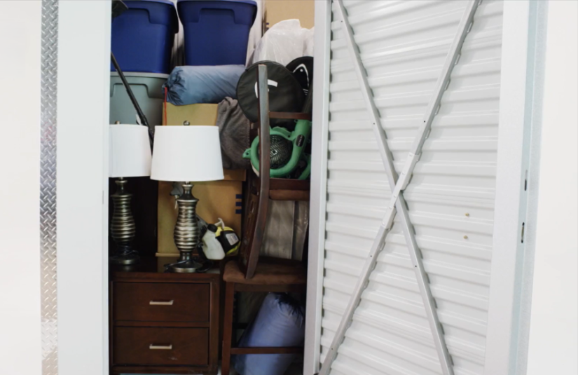 A view of the interior of a 5x5 storage unit, which is full of furniture, bins, and table lamps.