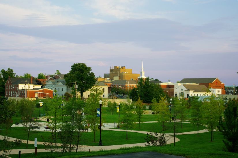 This image depicts a serene view of Cookeville, Tennessee, at sunset. In the foreground, a lush green park is visible, featuring winding walking paths, lampposts, and benches. The background showcases a mix of charming buildings, including red-brick and light-colored structures, with a prominent white church steeple standing out against the skyline. The scene is bathed in soft evening light, with a pastel-colored sky completing the peaceful ambiance.