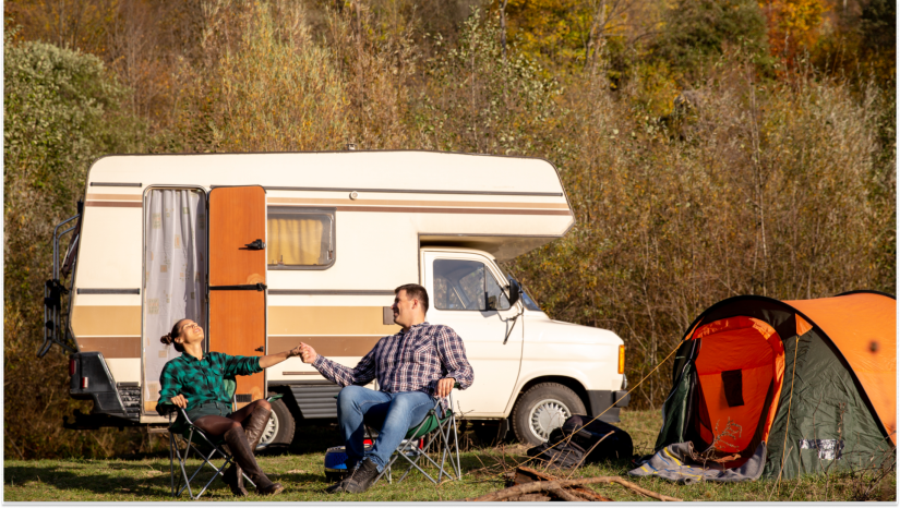 couple sitting outside of an rv with an orange tent to their right with bushes behind them