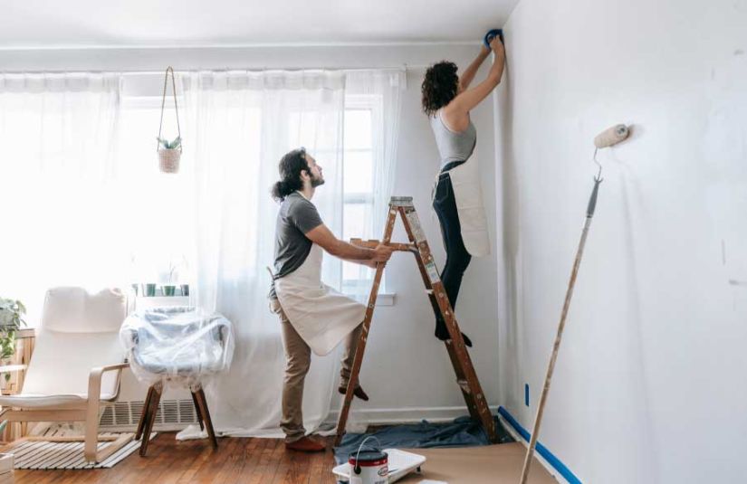 a man holding a ladder while a women tapes the edge of the wall before painting