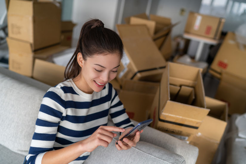 A woman surrounded by boxes uses her phone to reserve a storage unit online.