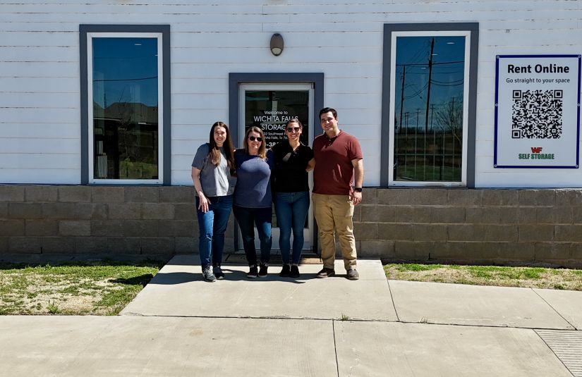 Four adults, two men and two women, stand smiling in front of a white building with "Wichita Falls Self Storage" over the door. A "Rent Online" sign with a QR code is visible on the right side of the building.