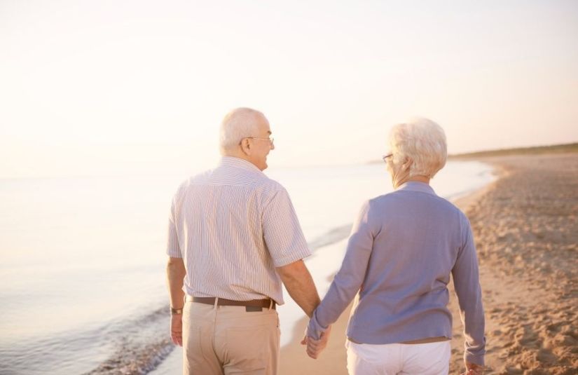 A retired couple holding hands on a beach at sunset.