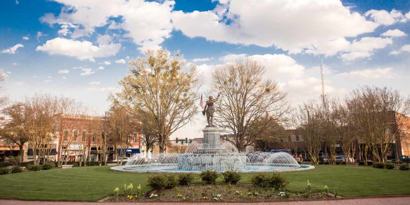 This image features Lafayette Square in LaGrange, Georgia, showcasing a vibrant public space with a beautiful fountain as its centerpiece. The fountain is surrounded by well-maintained green lawns, colorful flowers, and a statue of the Marquis de Lafayette standing proudly atop the structure. The background includes charming historic buildings, tree-lined streets, and a clear blue sky with scattered clouds, creating a welcoming and picturesque downtown scene.