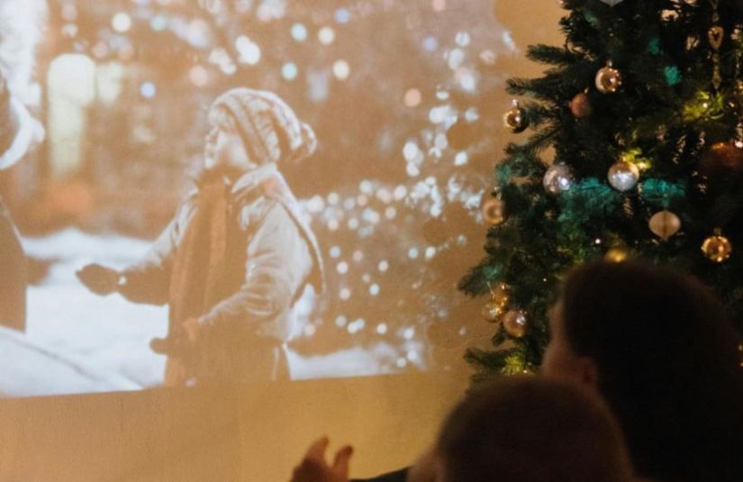 Two people watch a Christmas movie projected on a wall behind a decorated Christmas tree.