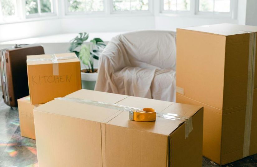A collection of moving boxes, labeled with specific rooms and sealed with packing tapes. A chair covered in a dust cloth and a suitcase sit behind the boxes.