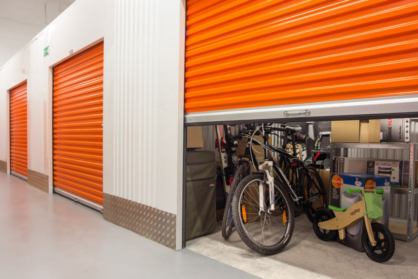 Self-storage unit with an open orange door revealing bicycles, boxes, and household items inside.