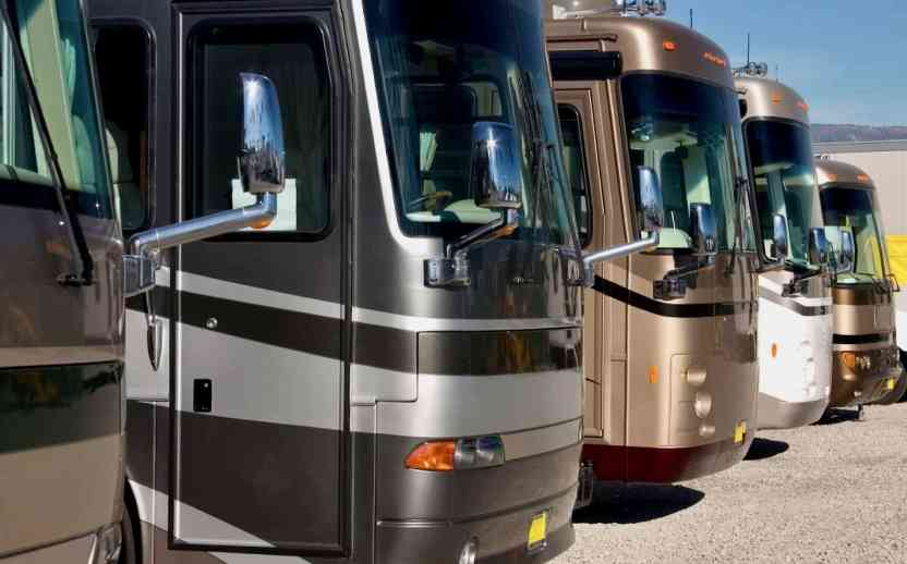 A row of Class A RVs parked side-by-side on a gravel lot at a secure vehicle storage facility.