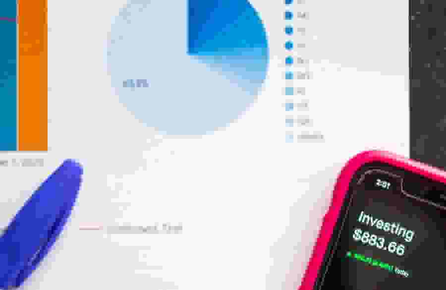 A phone with a screen reading "Investing: $883.66" rests on a paper with graphs and charts. A pen rests nearby to annotate.
