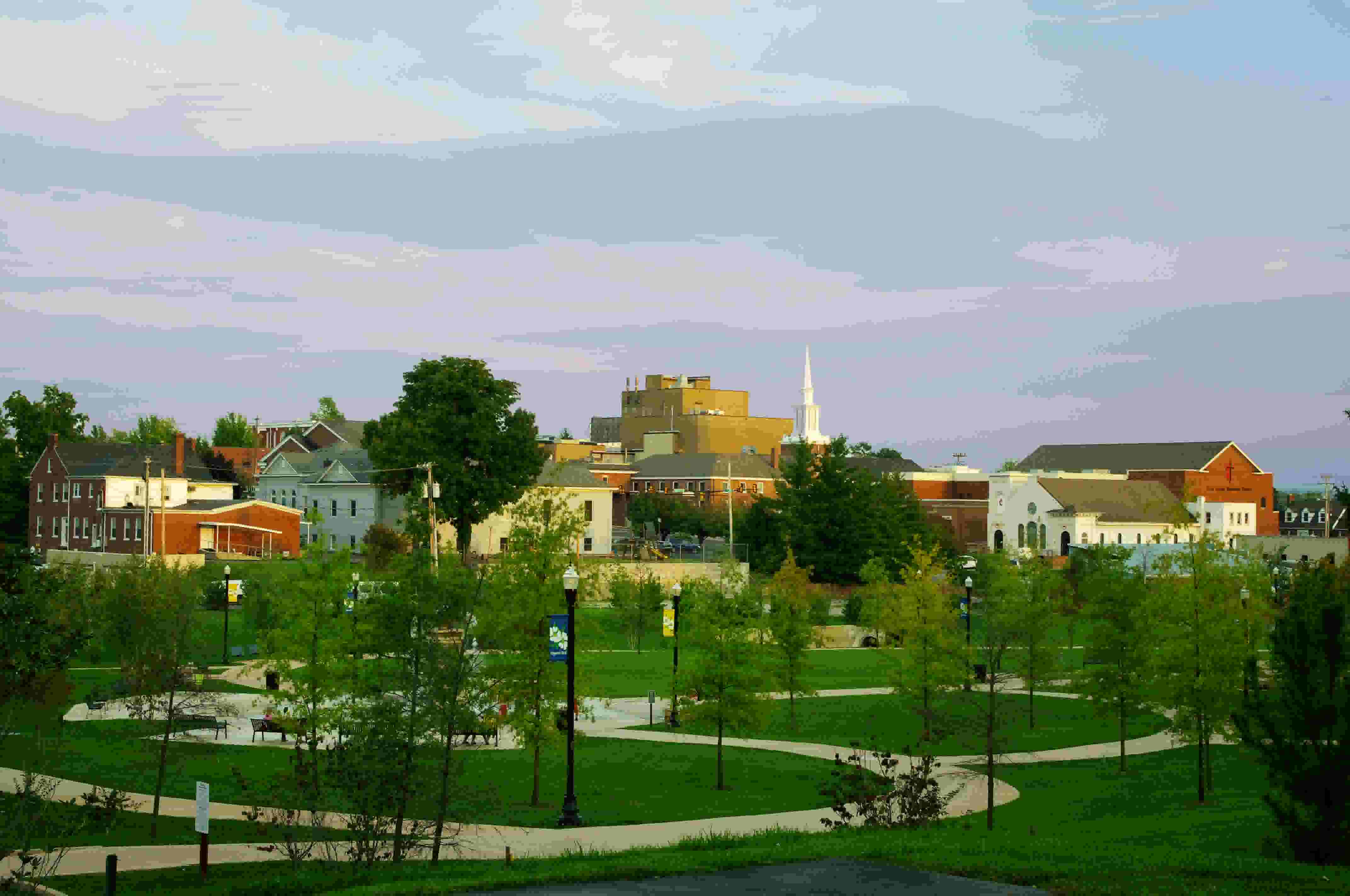 This image depicts a serene view of Cookeville, Tennessee, at sunset. In the foreground, a lush green park is visible, featuring winding walking paths, lampposts, and benches. The background showcases a mix of charming buildings, including red-brick and light-colored structures, with a prominent white church steeple standing out against the skyline. The scene is bathed in soft evening light, with a pastel-colored sky completing the peaceful ambiance.