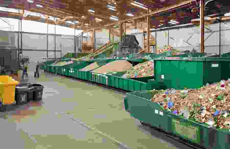This image shows a recycling plant actively in use, the foreground has green bins full of waste and the background has workers walking around the facility.