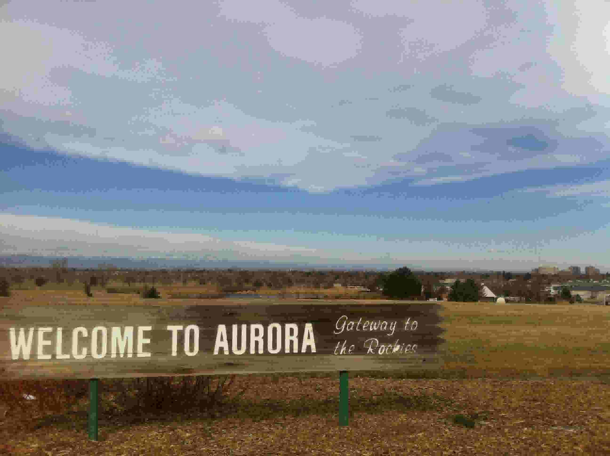 A photograph of a sign that reads "WELCOME TO AURORA: Gateway to the Rockies." The city of Aurora and the mountains are visible in the background.
