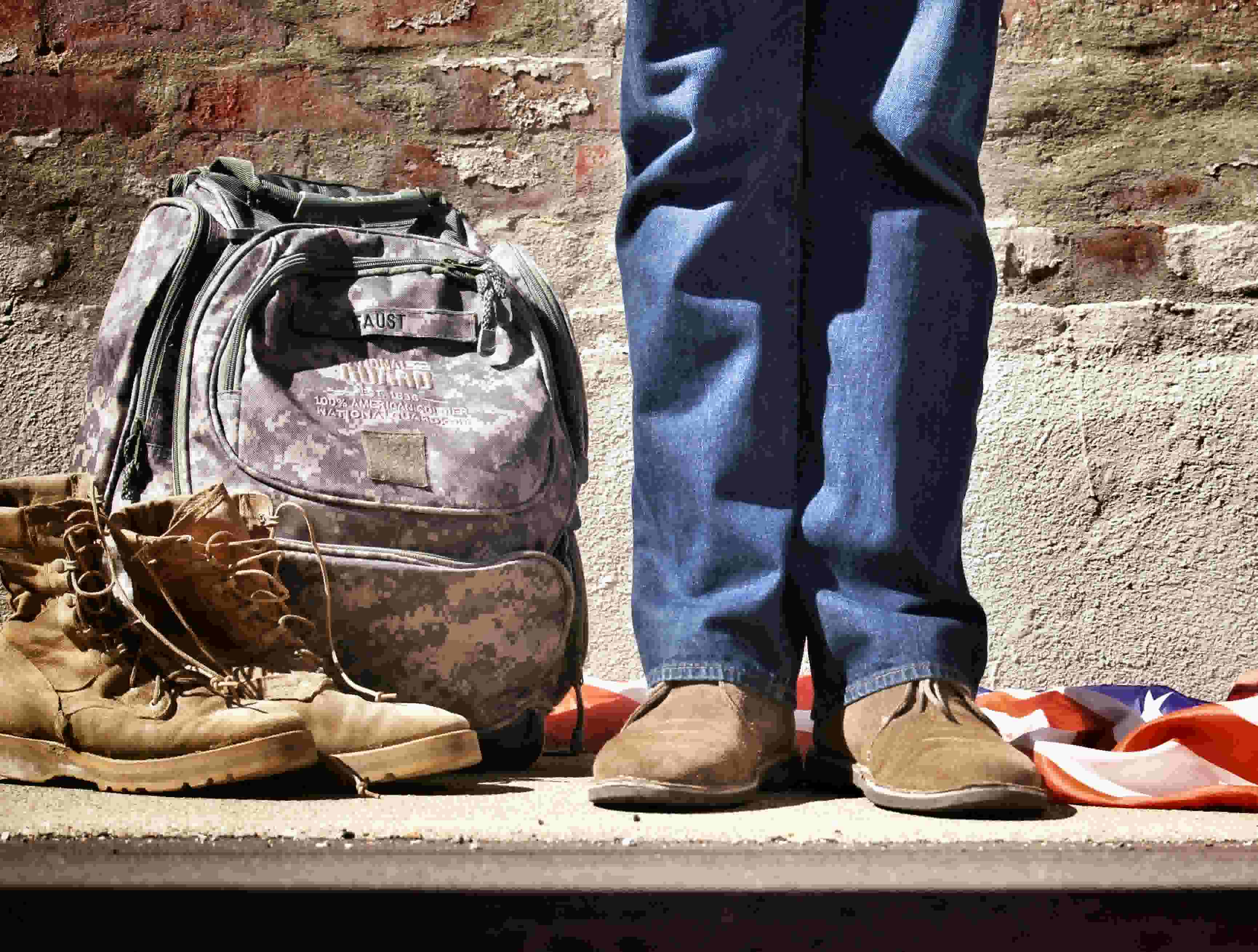 A person stands near a pair of boots, a military backpack, and an American flag.