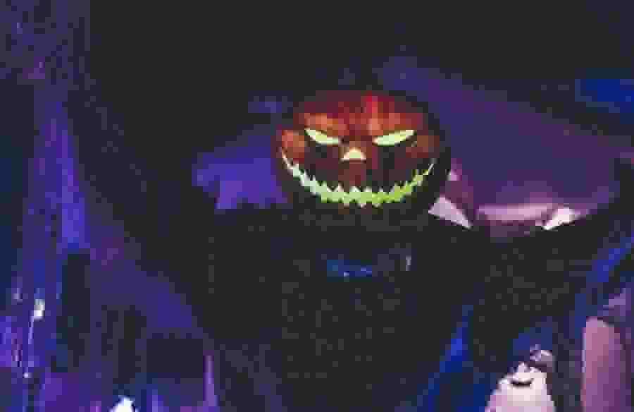 A person in a spooky costume with a glowing jack o'lantern head looms towards the camera.