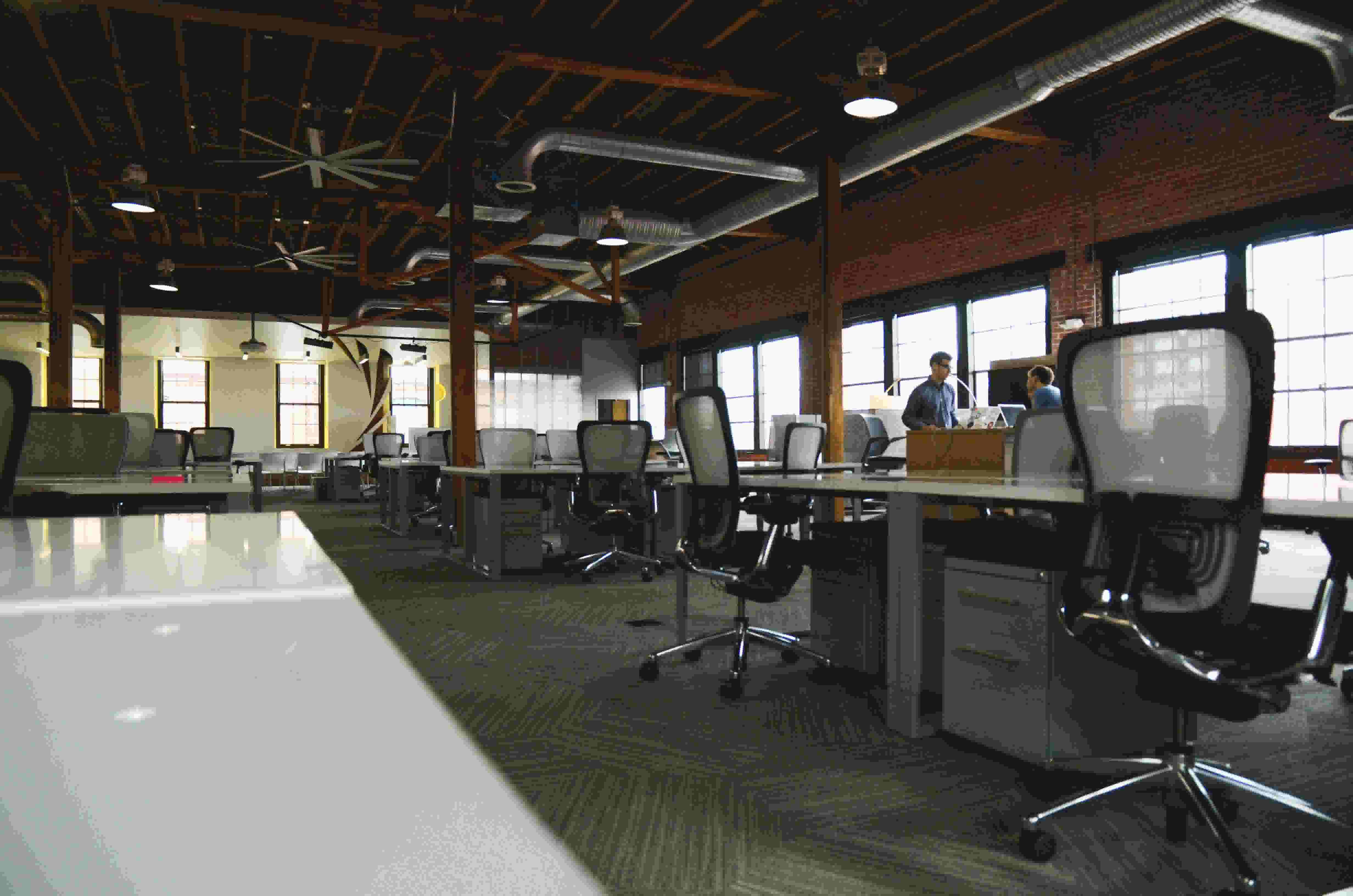 An office space. Rolling chairs sit at desks. In the background, two coworkers have a conversation.