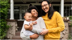 family in various clothing colors smiling with a baby standing outside of a home with green plants