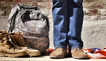 A person stands near a pair of boots, a military backpack, and an American flag.