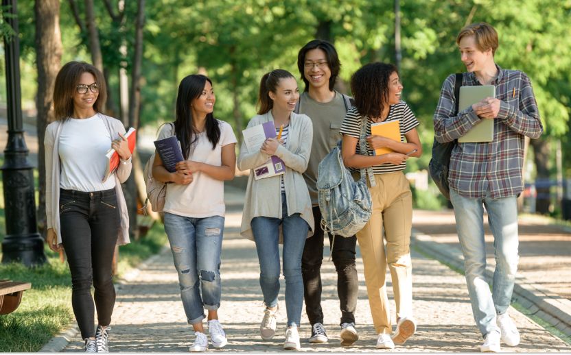 students in various colored clothing walking through a paved park holding various study material