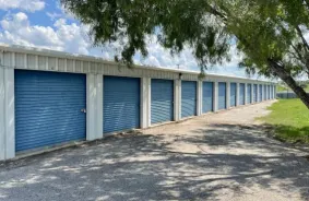 Outdoor storage unit with white roll up doors and a tree in front on a gravel road
