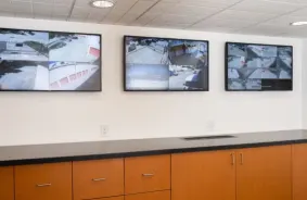 Three wall-mounted screens show video surveillance footage of the facility.